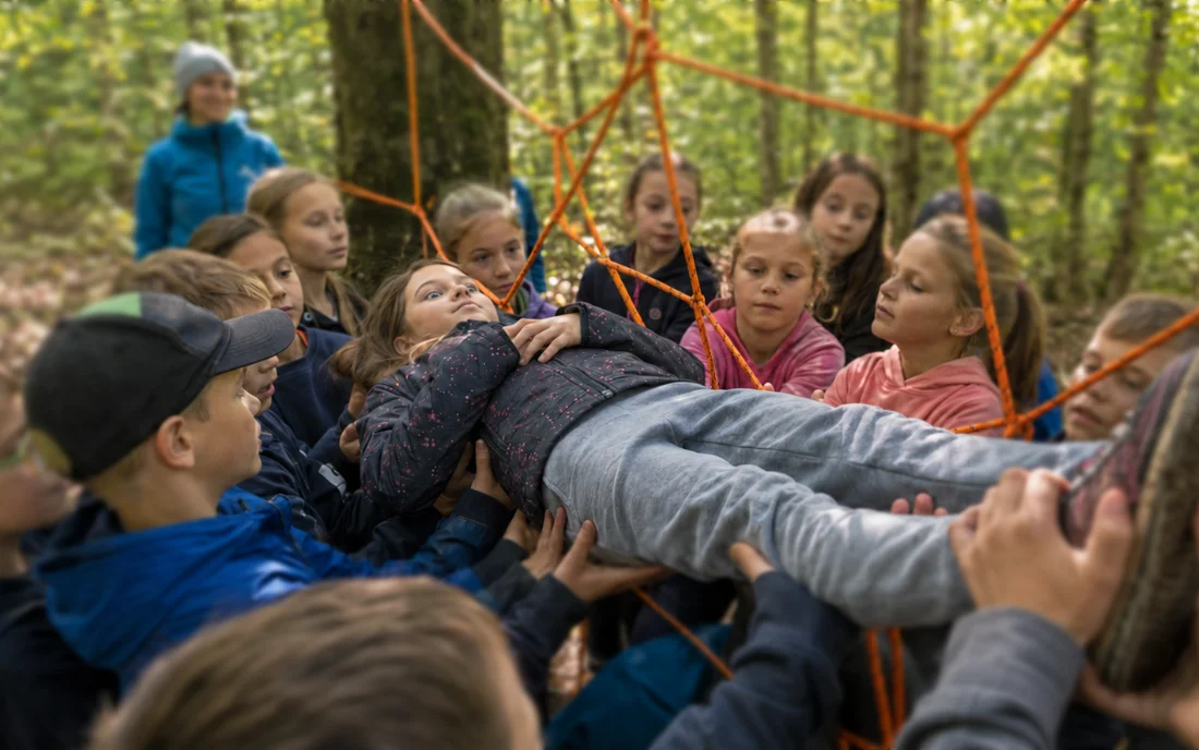 Grundschulkinder tragen eine Mitschülerin gemeinsam durch ein Seilnetz im Wald.